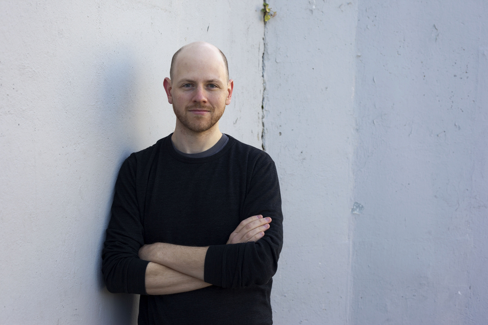 Dan Moren smiling with arms crossed, leaning against a white concrete wall.