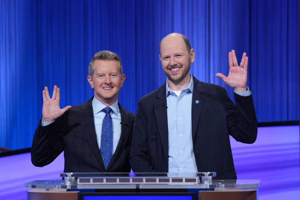 Dan standing with Ken Jennings behind a Jeopardy! podium, both of them doing a Vulcan salute.